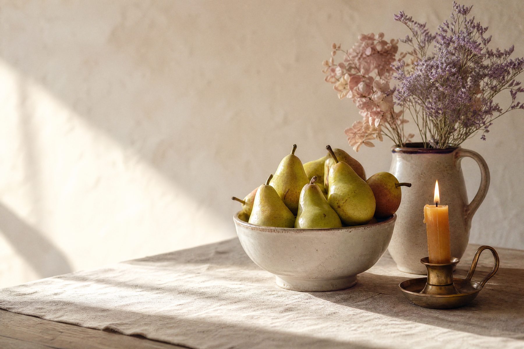 A ceramic bowl of pears, a jug of dried flowers and a candle on a sunlit table